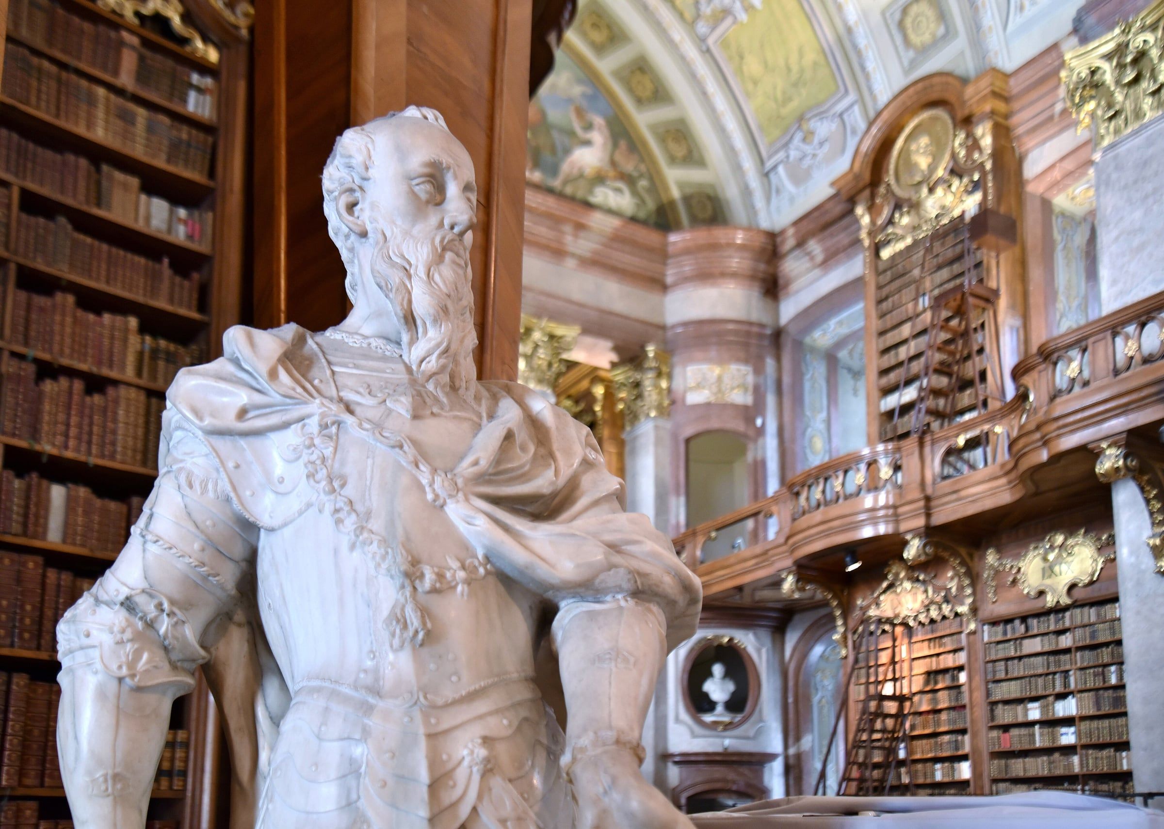 A marble statue of an armoured figure stands before the Baroque bookshelves and gilded galleries of the Austrian National Library State Hall.