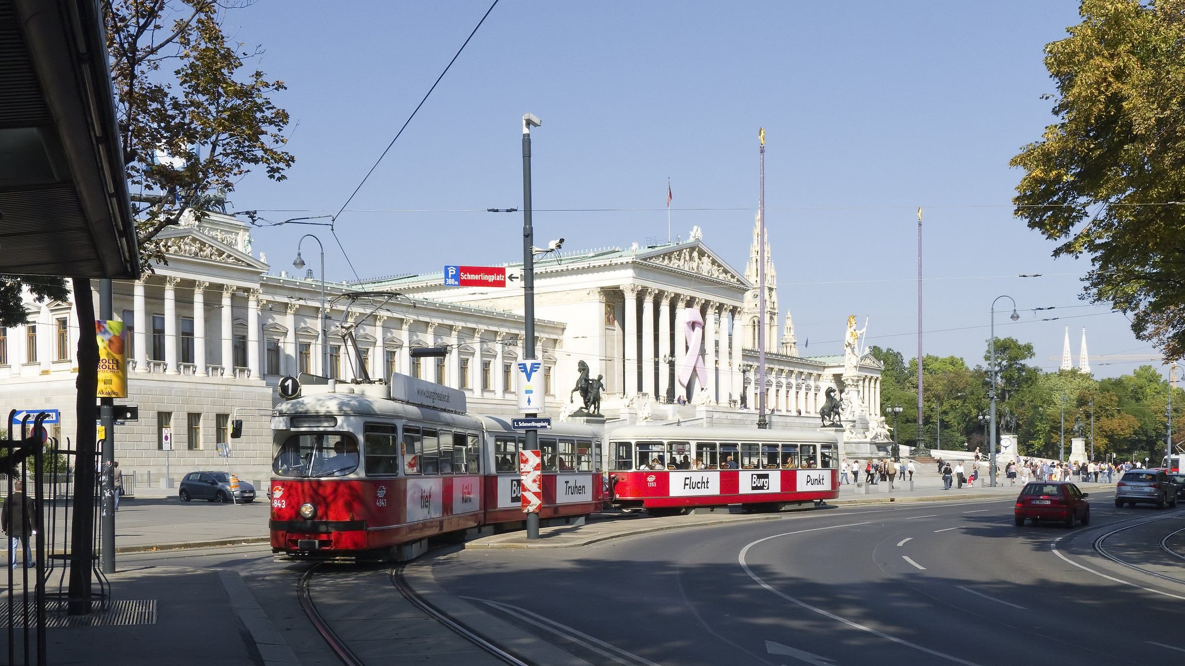 A red and white tram passes a neoclassical columned building and its statues on the Ringstrasse under a clear blue sky.